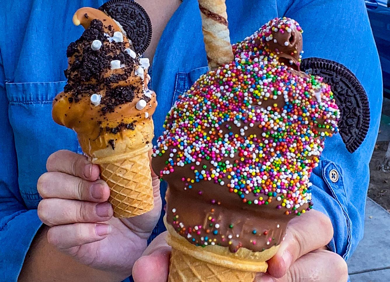 Two chocolate dipped ice cream cones topped with sprinkles and Oreo, held up outside Dipped Ice Cream